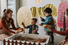 Imagem de uma mãe utilizando a parte da rotina do bebê para brincar, com dois filhos na idade entre 1 e 3 anos. Na frente das crianças um berço decorado e atrás, um quarto colorido com decorações.