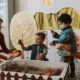 Imagem de uma mãe utilizando a parte da rotina do bebê para brincar, com dois filhos na idade entre 1 e 3 anos. Na frente das crianças um berço decorado e atrás, um quarto colorido com decorações.